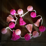 Alternating bright pink and light pink pompoms and tassels on a white crocheted string against a wooden floor
